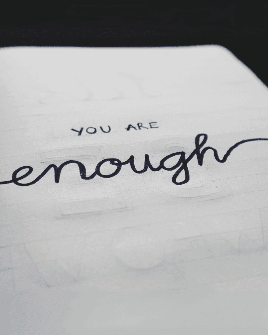 Close-up of an open journal with the handwritten phrase “You are enough” on the page, next to a pen on a cozy desk or bed setting.