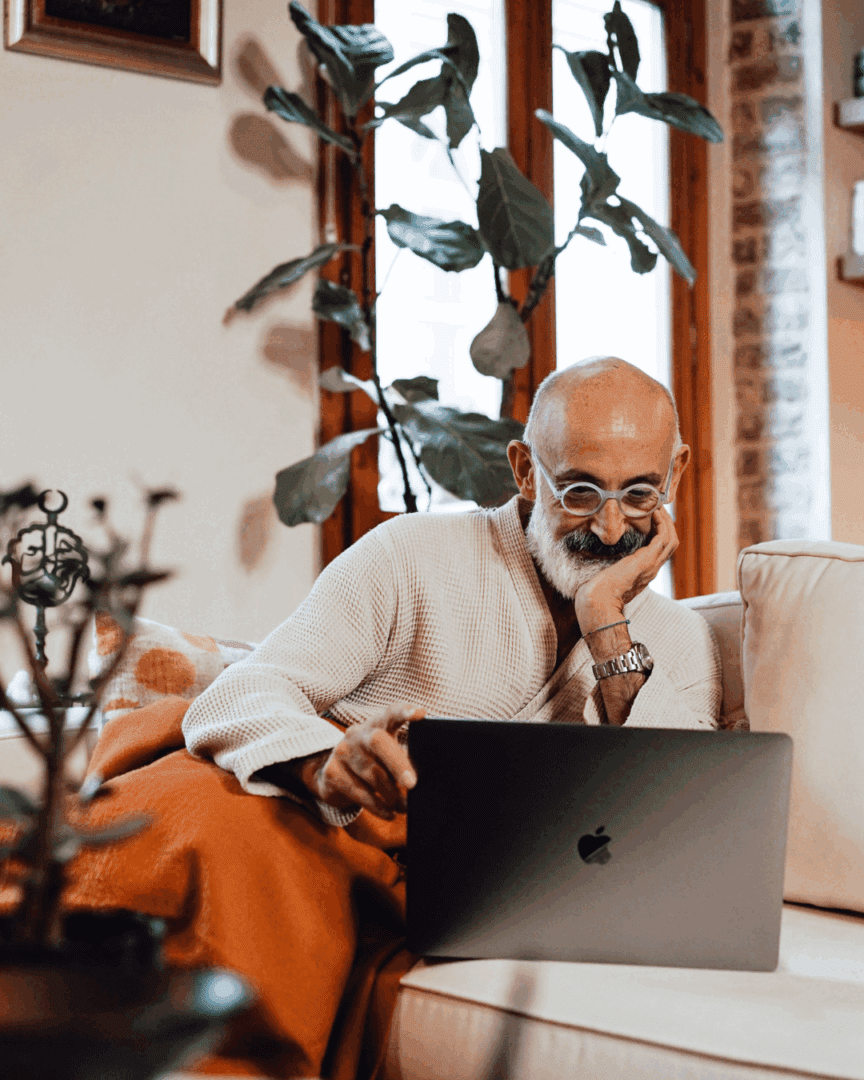 An older man in glasses sits on a couch with a laptop, wrapped in a blanket, reflecting quietly capturing the question many ask in therapy, why do I feel broken.