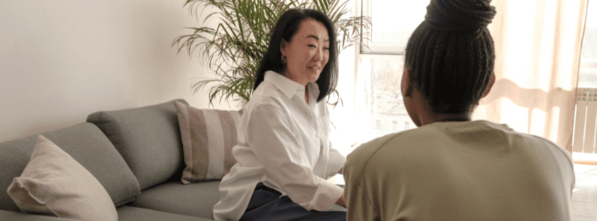 A woman speaks with another in a sunlit living room, showing the supportive space therapy provides when clients wonder why do I feel broken.