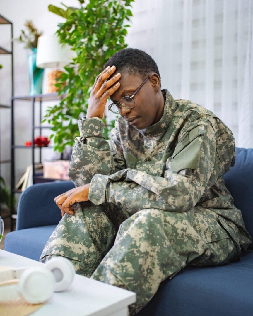 A Black person in military camouflage sits on a couch with their hand against their forehead, expressing emotional strain often tied to helper burnout, stress, or transition challenges among veterans.