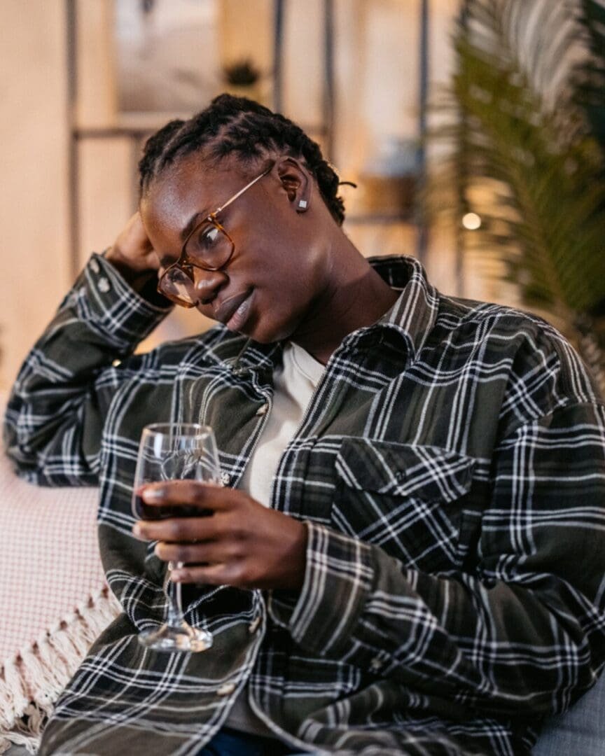 A non-binary person sits on a couch holding a glass of wine with their head resting on their hand, appearing overwhelmed or sad during the holidays, reflecting holiday grief and emotional fatigue.