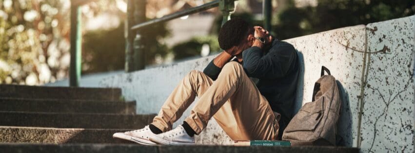 Young man sitting alone on outdoor concrete steps with his head down and hands covering his neck, backpack beside him, appearing overwhelmed or depressed in Atlanta GA near 30339 and Savannah 31401.