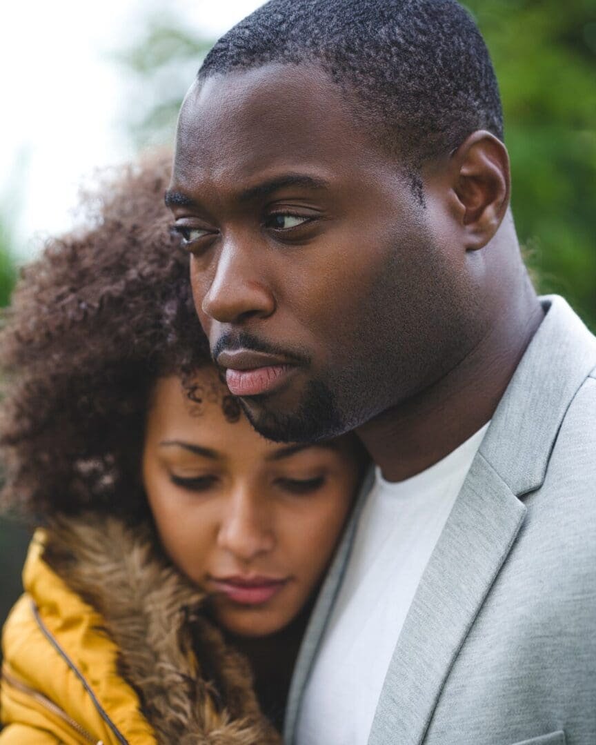 Close-up of a Black couple standing outdoors; the man looks serious and protective while the woman rests her head against his chest, both appearing thoughtful and emotionally connected in Atlanta GA near 30327 and Savannah 31401.