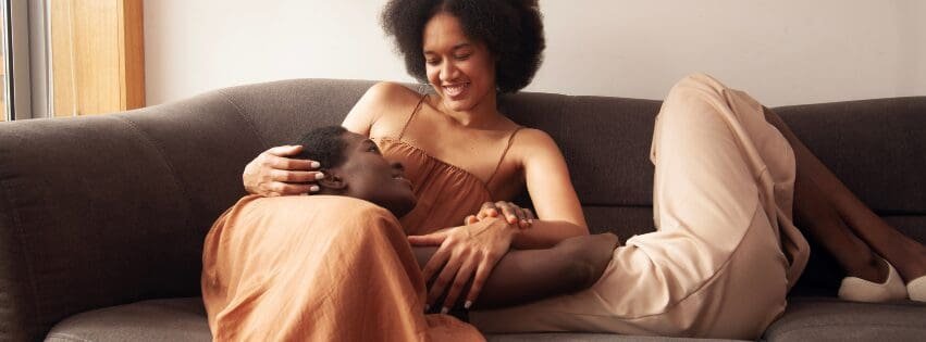 Black queer couple relaxing on a couch at home; the woman smiles gently while the man rests his head on her lap, showing affection and emotional closeness in Atlanta GA near 30327 and Savannah 31401.