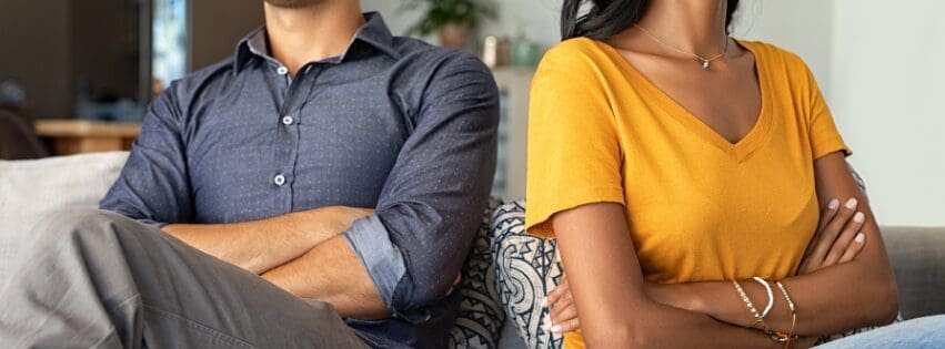 Two adults sit on a couch with their bodies turned away from each other and arms crossed, showing emotional distance and unresolved conflict often addressed in couples therapy and relationship counseling in Atlanta GA near 30308 and 30312.