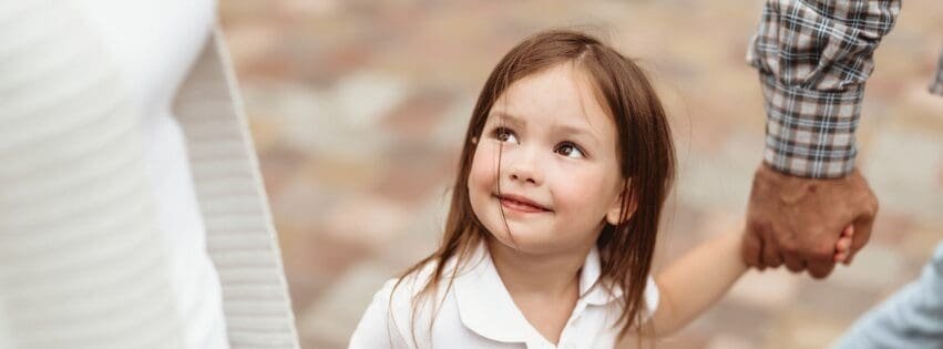 A young child holds the hands of two adults while walking outdoors in Georgia, looking up at one caregiver with a curious, attentive expression.