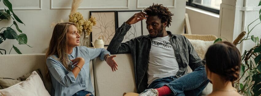 Three adults sit in a therapy room having a serious conversation. Two people gesture while talking on a couch, and a third person listens nearby, suggesting a tense or emotionally complex discussion.
